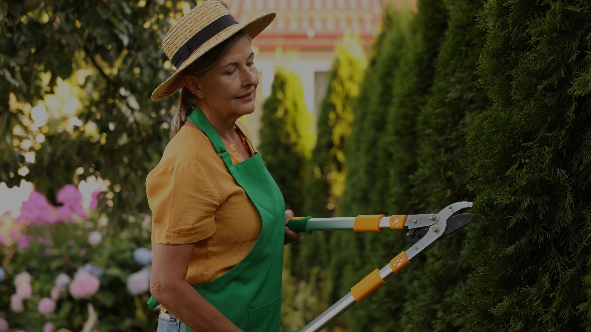Person in a garden wearing a straw hat and green apron, holding gardening shears.