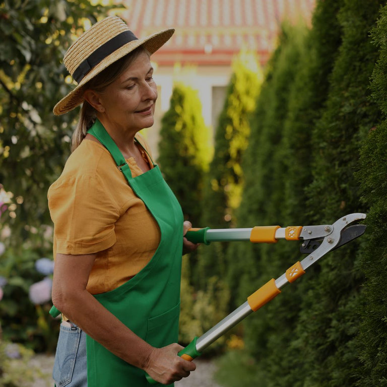 Person in a garden wearing a straw hat and green apron, holding gardening shears.