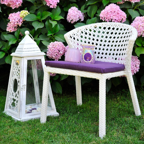 White wicker chair with purple cushion and decorative pillow, accompanied by a white lantern and pink flowers.